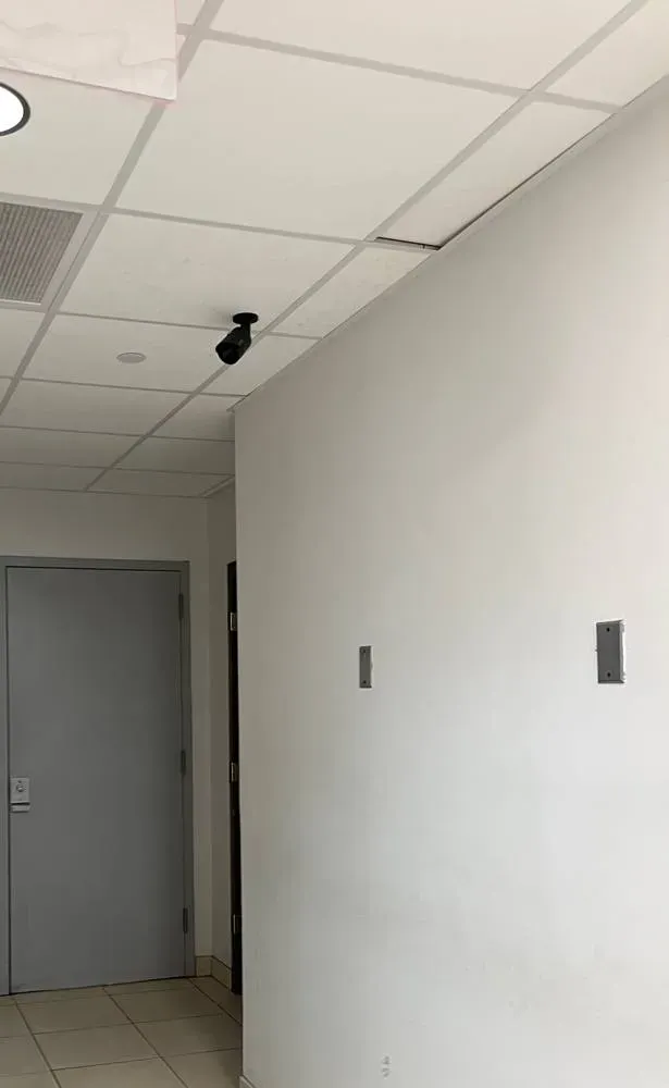 Bullet camera above a hallway door — NYC corridor camera reference Wireless camera installation NYC reference photo: a black bullet camera on a short mount at the edge of a white drop-ceiling grid, above a grey hallway door, with plain white walls and two small metal wall plates