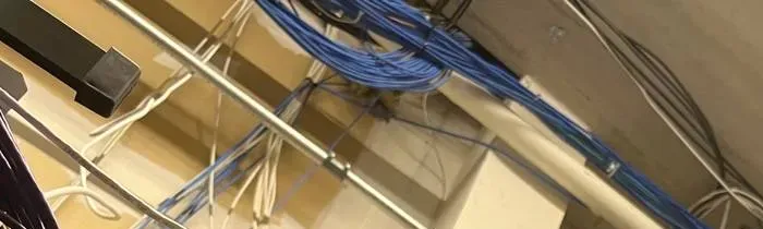 Network cables bundled along a ceiling pipe — NYC PoE camera cabling reference Wireless camera installation NYC reference photo: bundles of blue and white network cables running along a metal pipe and ceiling structure, with a dark bracket at the left edge