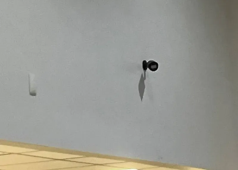 Small camera high on a bare wall — NYC wireless camera mounting reference Wireless camera installation NYC reference photo: a small black camera on a short mount high on a bare pale wall, casting a long shadow below it, with a white wall plate to the left